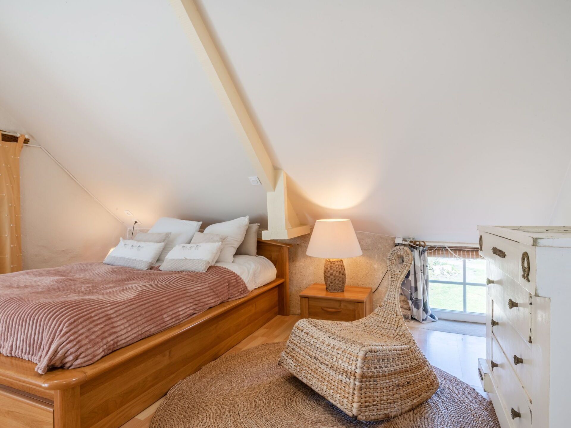Een slaapkamer met een houten bed, een geweven poef, een nachtkastje met lamp en een witte dressoir.