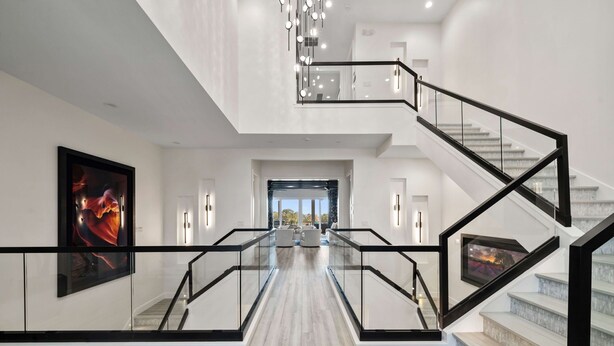 Un interior moderno con una escalera de barandilla de cristal, una obra de arte grande enmarcadas en la pared y vista a un área de estar.