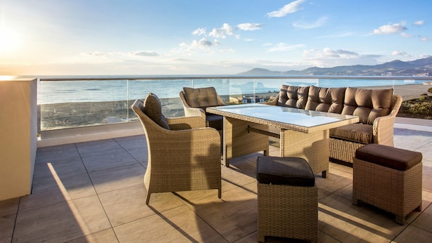 A balcony with wicker furniture, a glass table, and a view of the sea and mountains.