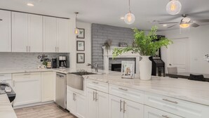 Une cuisine moderne avec des meubles blancs, un plan de travail en marbre, un lave-vaisselle intégré et un îlot central. On y trouve des luminaires suspendus, une cheminée et une machine à café sur le plan de travail.
