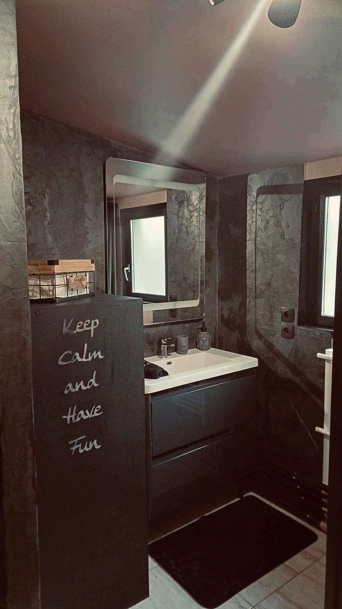 Une salle de bain avec un mur foncé à la texture marquée, un lavabo blanc et un miroir au-dessus. Un tapis noir est posé sur le sol et un message est écrit sur le mur : « Restez calme et amusez-vous. »