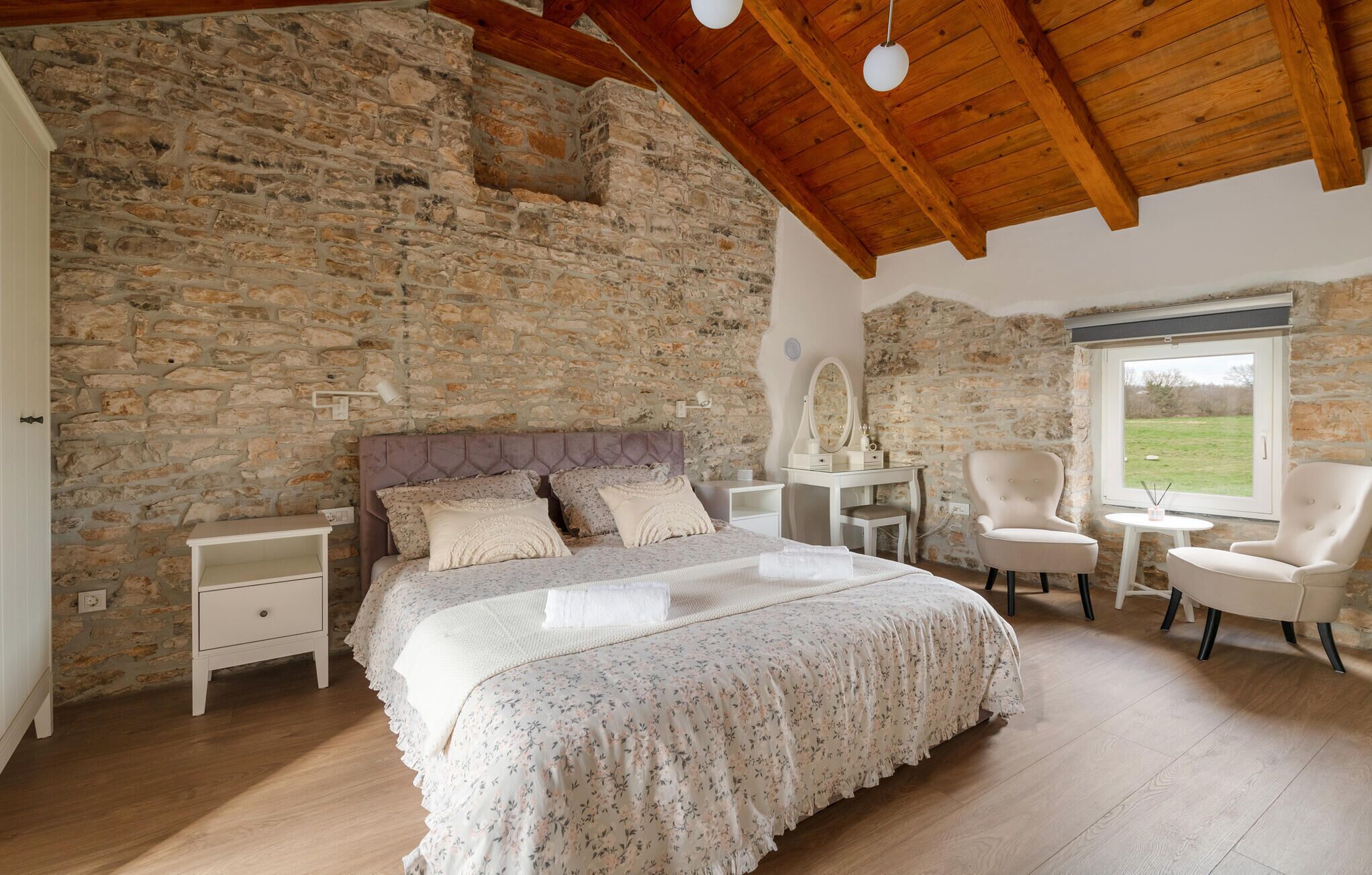 A bedroom with a stone wall, wooden ceiling beams, a bed with floral bedding, bedside tables, a chair, and a window with a view.