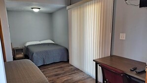 A small, compact room with a single bed, a desk, and a chair. There is a window with vertical blinds, a nightstand, and a wall socket. - The Weekender Unit #1 (Geneva-on-the-Lake)