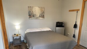 A bedroom with a bed, a bedside table with a lamp, a refrigerator, and a painting on the wall. - The Weekender Unit #2 (Geneva-on-the-Lake)