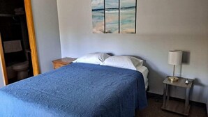 A bedroom with a bed, a nightstand, a lamp, and a painting on the wall. - The Weekender Unit #4 (Geneva-on-the-Lake)