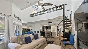 A spacious living area with a high ceiling, a modern staircase, and a view of the kitchen and dining area.