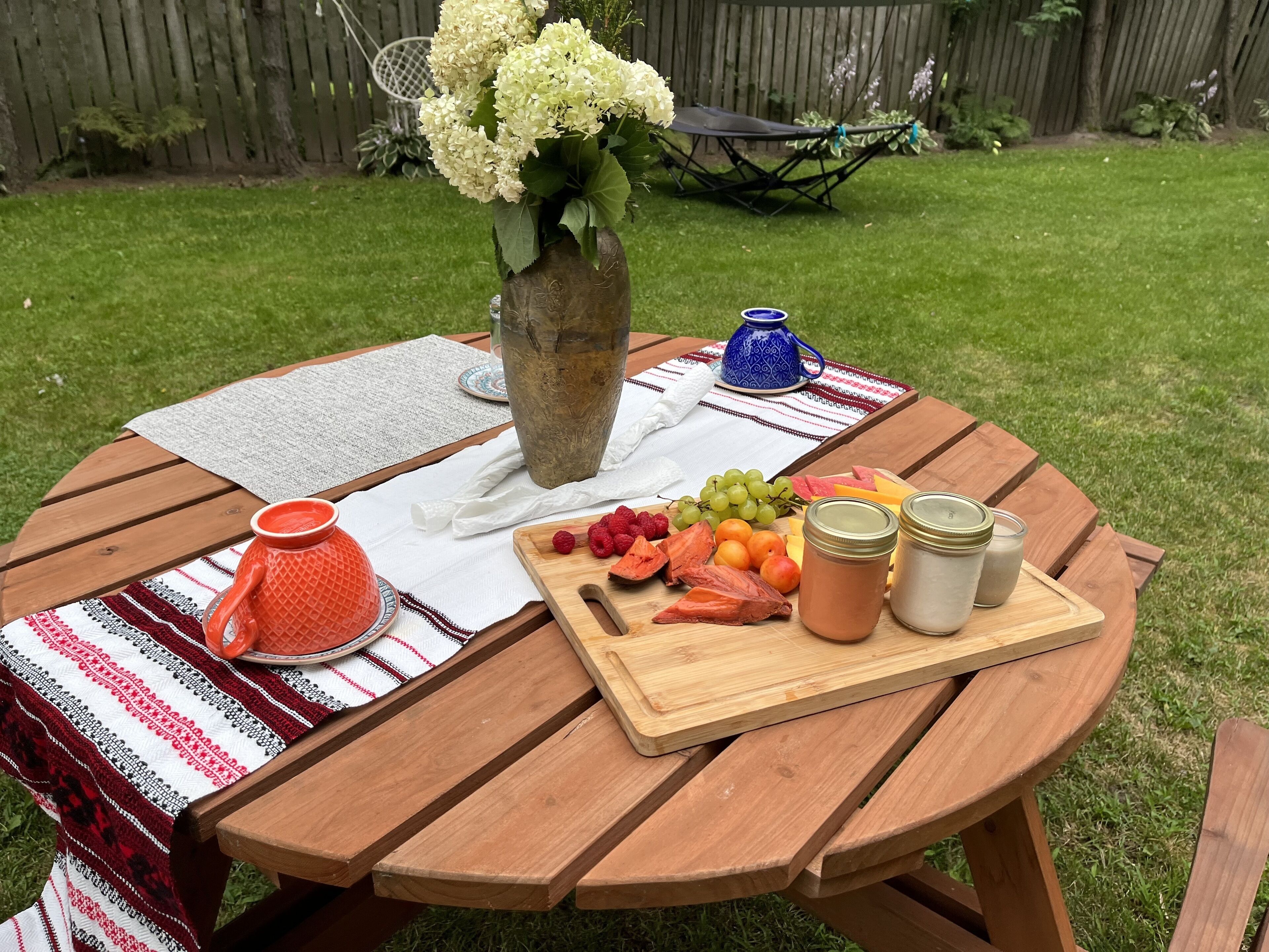 Uma mesa de piquenique de madeira com toalha branca, um vaso com flores, um jarro azul e uma tábua de madeira com frutas e potes.
