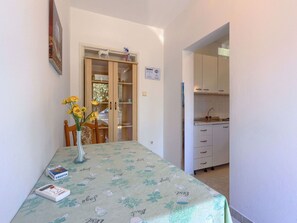 Un petit coin repas avec une table dressée pour deux, un vase rempli de fleurs jaunes et une télécommande posée sur la table. Au fond, on aperçoit un buffet en bois avec des portes en verre, et une partie de la cuisine est visible à travers une porte ouverte.