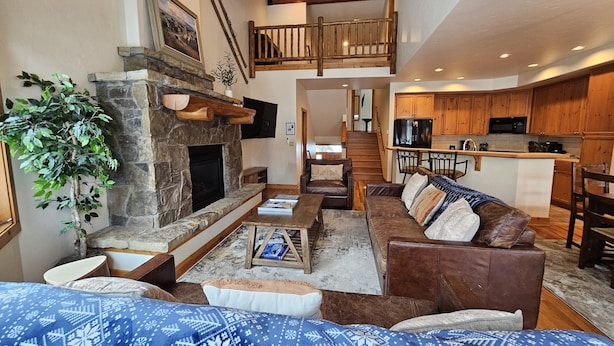 A spacious living area with a stone fireplace, a large sofa, and a dining table. A kitchen is visible in the background with wooden cabinets and a built-in microwave.
