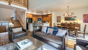 A spacious living area with a sofa, coffee table, and a dining table with chairs. A kitchen is visible in the background with wooden cabinets and a built-in oven.