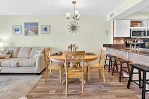 A dining area with a round table, wooden chairs, a chandelier, and a kitchen in the background.
