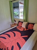 Un dormitorio con cama, cortinas verdes, ventana con vistas a la vegetación y mesita de noche con lámpara.