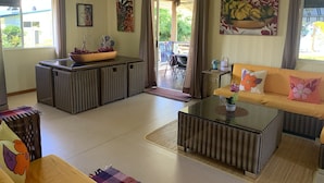 Amplia sala de estar con sofá, mesa de centro y mesa de comedor. Se aprecia una ventana con cortinas, un cuadro en la pared y vistas al exterior.