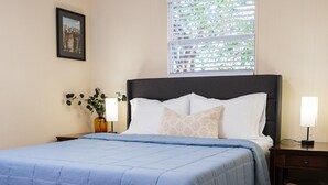 A bedroom with a bed, a nightstand, a lamp, a framed picture on the wall, and a window with blinds.