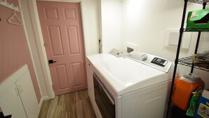 A laundry room with a washing machine, a dryer, a pink door, and a shelf with cleaning supplies.