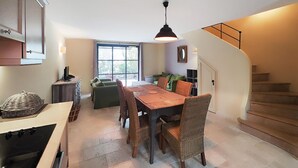 Un coin repas avec une table en bois et des chaises en osier, une cuisine équipée d’une cuisinière et un escalier menant à un étage supérieur.