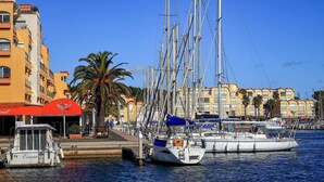 Un port de plaisance où sont amarrés de nombreux voiliers, un restaurant avec un auvent rouge et une rangée de bâtiments jaunes dotés de balcons.