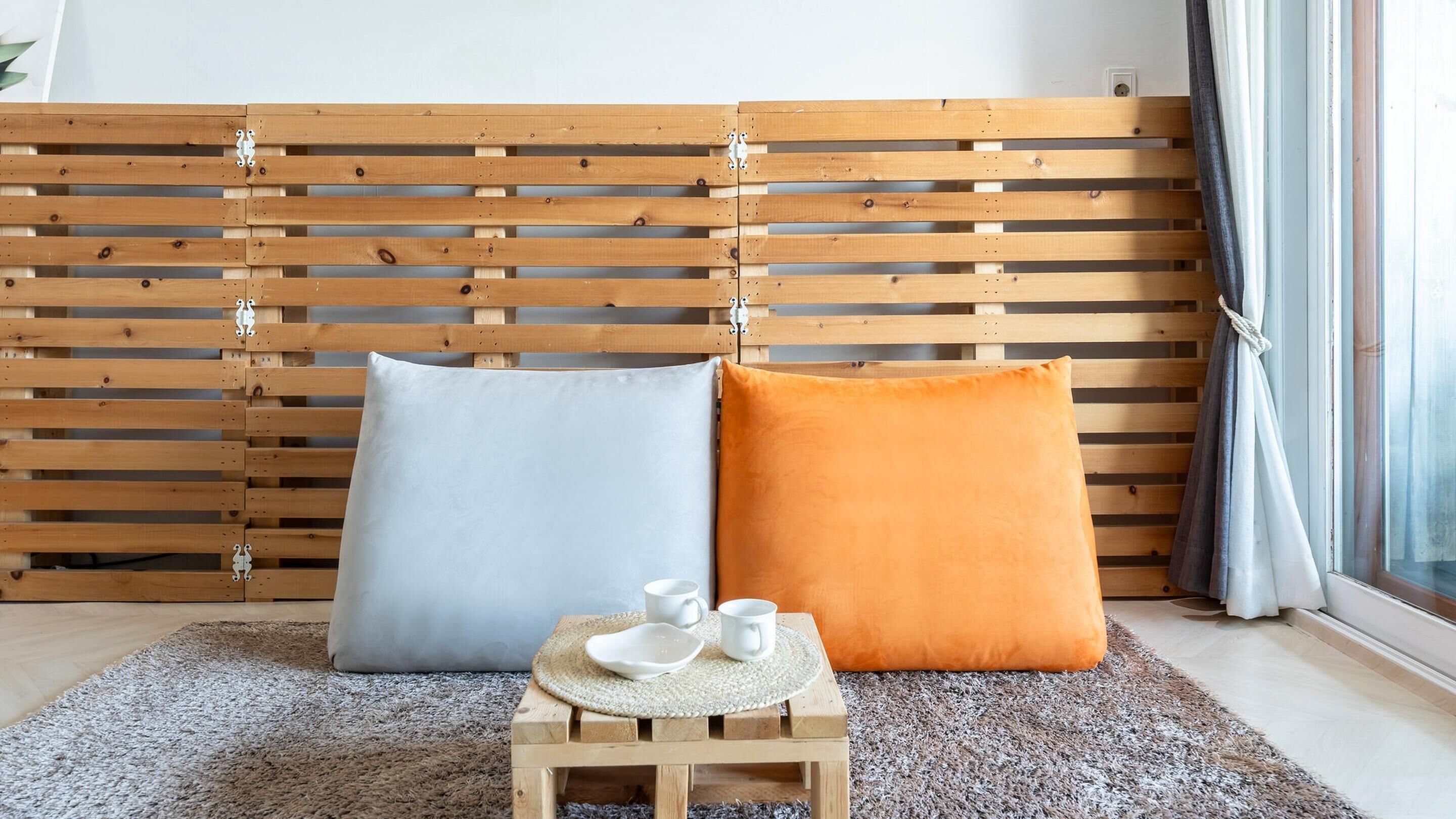 Una habitación con cabecera de madera con listones, dos almohadas (una azul claro y una naranja), una mesita de madera con dos tazas blancas y una alfombra con textura en el suelo.