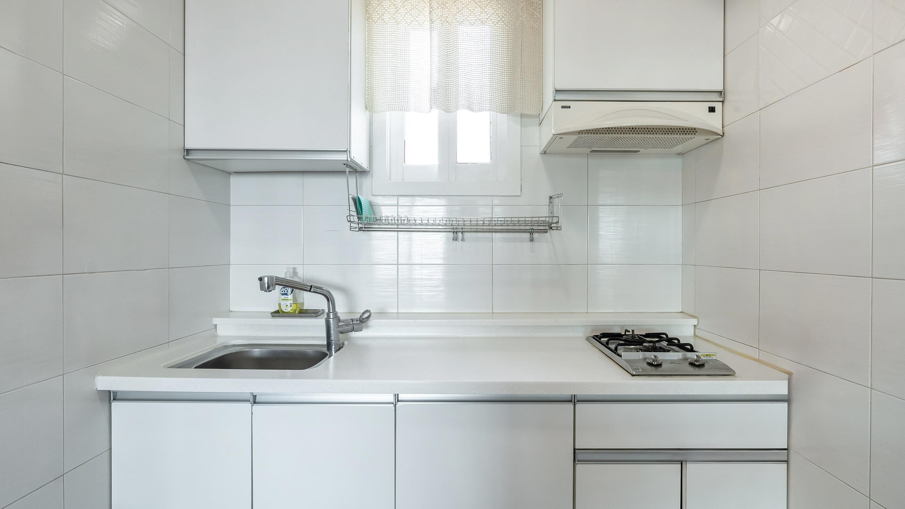 Una cocina moderna con gabinetes blancos, un lavamanos de acero inoxidable, una estufa a gas y una ventana con cortinas traslúcidas.