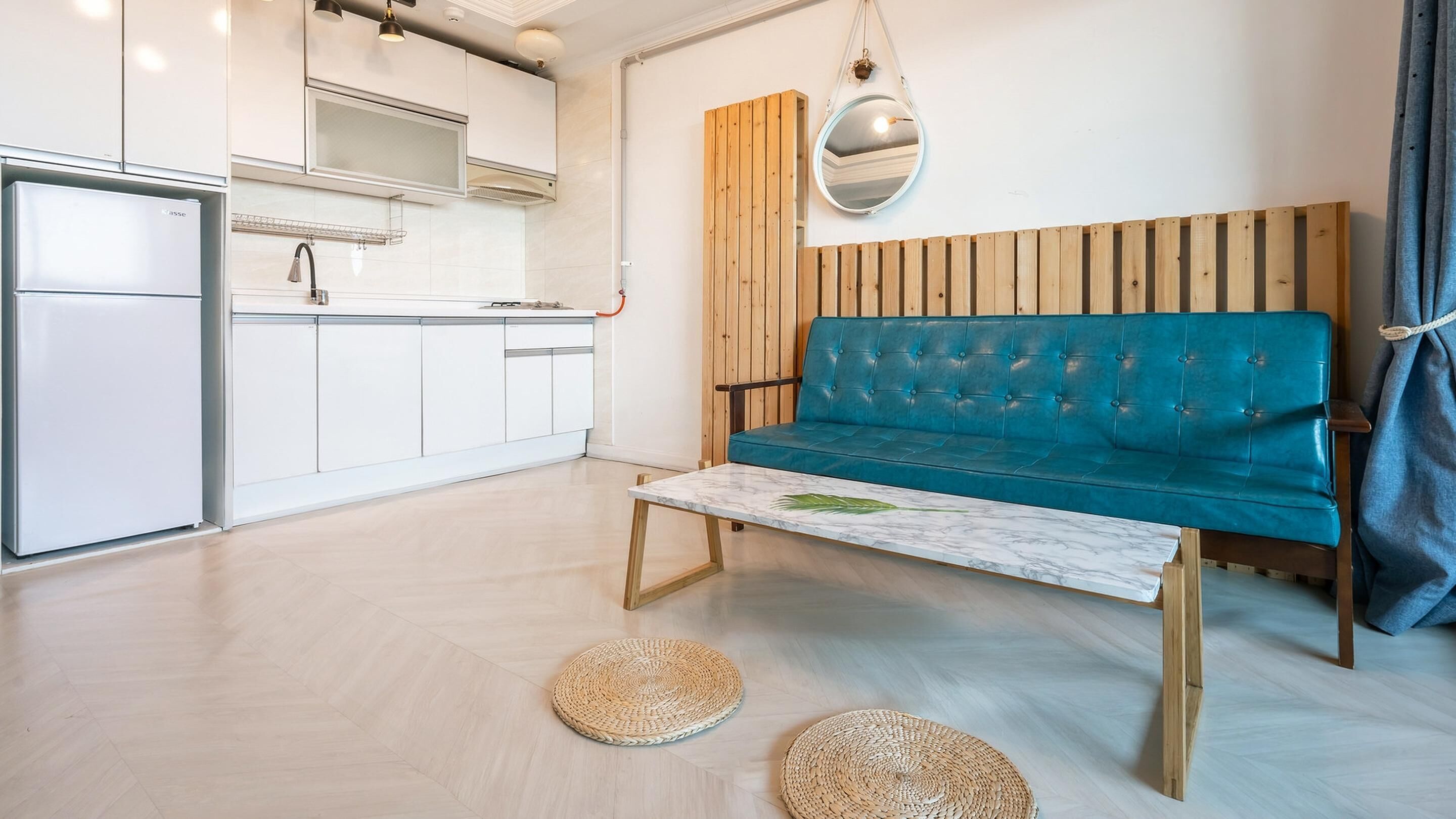 A modern living space with a kitchen, a teal sofa, a marble coffee table, and woven mats on the floor.