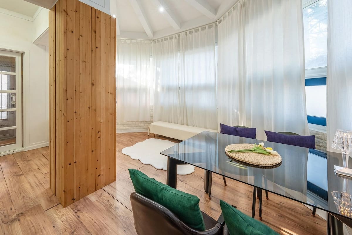 A modern dining area with a glass table, wooden panel wall, and a view of the outdoors.