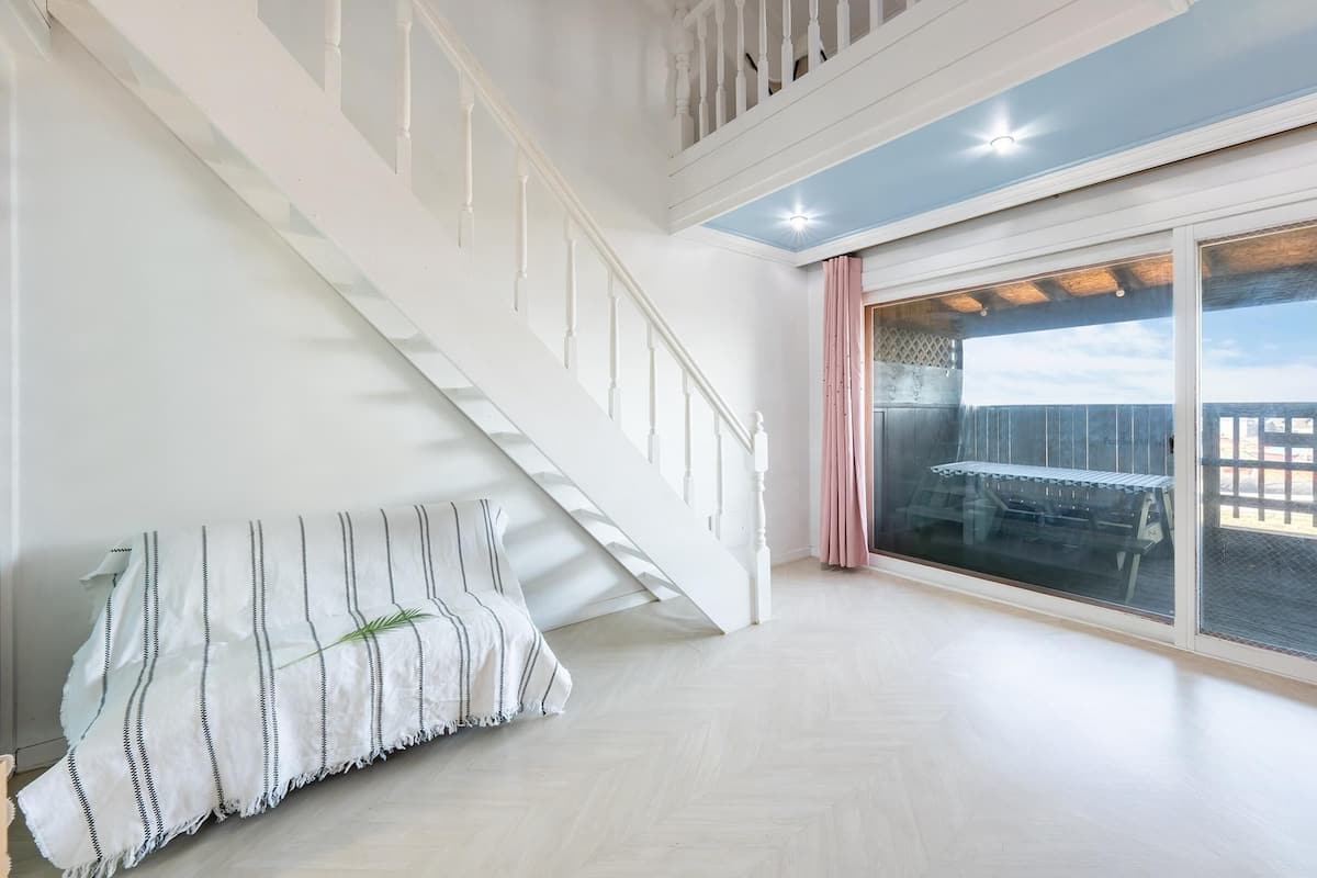 A spacious room with a wooden floor, a white staircase, and a large sliding glass door leading to an outdoor deck.