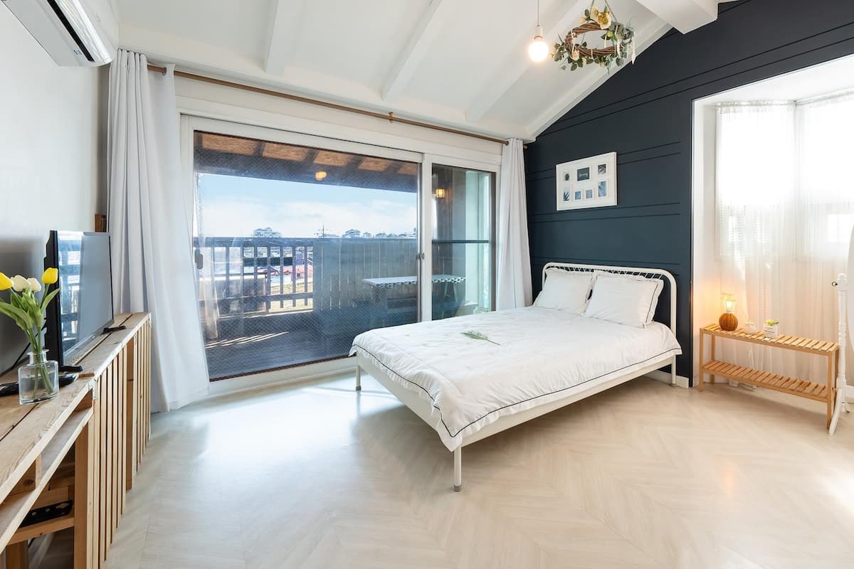 A bedroom with a bed, a bedside table with a lamp, a mirror, and a view of the outside through a sliding door.