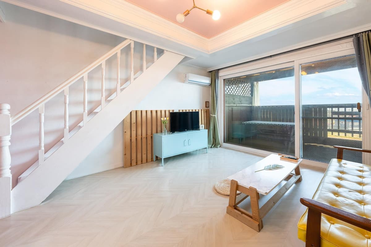 A spacious living room with a staircase, a TV on a wooden cabinet, a yellow sofa, and a large sliding glass door leading to an outdoor area.