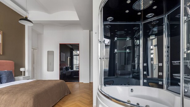 A modern bedroom with a large bed, a glass-enclosed jacuzzi, and a view of the adjacent room.