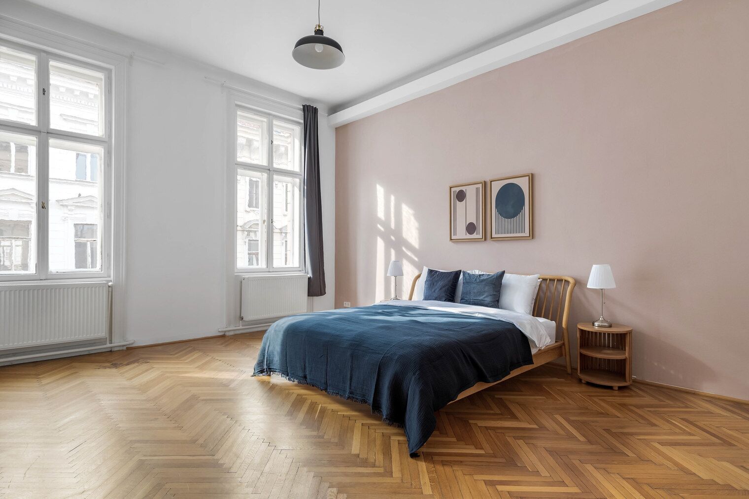 Ein Schlafzimmer mit Holzdielen im Fischgrätmuster, einem Bett mit blauer Bettdecke, einem Nachttisch aus Holz mit Lampe sowie zwei an der Wand befestigten abstrakten Kunstwerken.