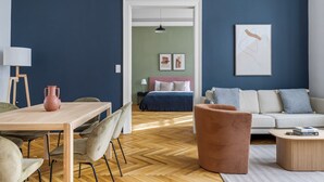 A modern living room with a wooden dining table, a sofa, and a coffee table. A bed with a green wall and a pink headboard is visible in the background.