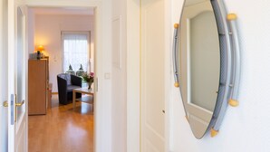 A hallway with a wooden floor leading to a room with a sofa and a table. A mirror with a decorative frame is mounted on the wall. - Apartment Kluge by Interhome (Zinnowitz)