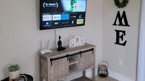 A living room with a flat-screen TV mounted on the wall, a wooden console table with decorative items, a small black side table with a plant, and a wall with the word "HOME" spelled out.