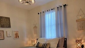 A cozy bedroom with a bed, a bedside table, a hanging light fixture, and a window with curtains.