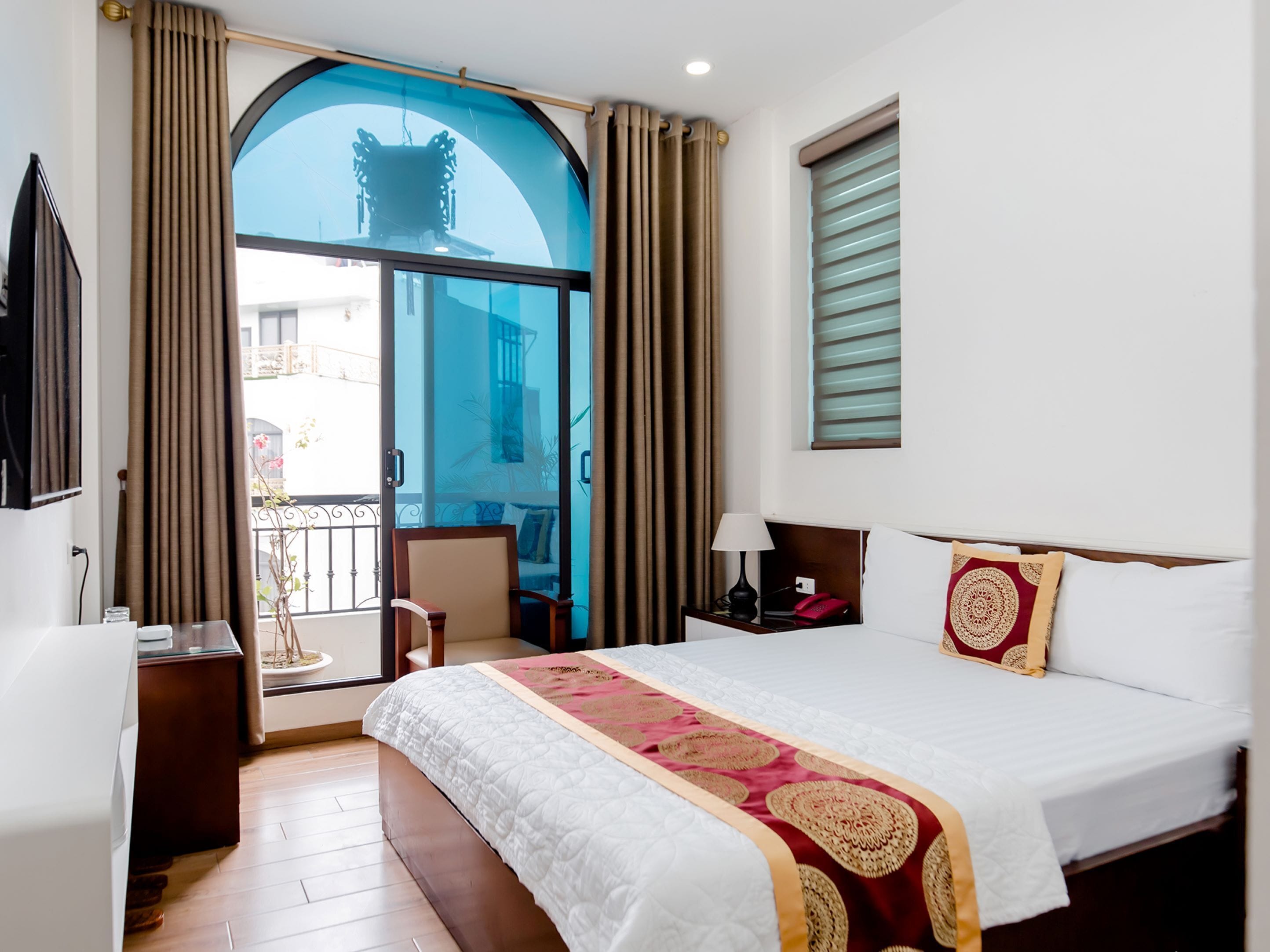 A hotel room with a large bed, a desk, a chair, and a balcony with a view of the city.