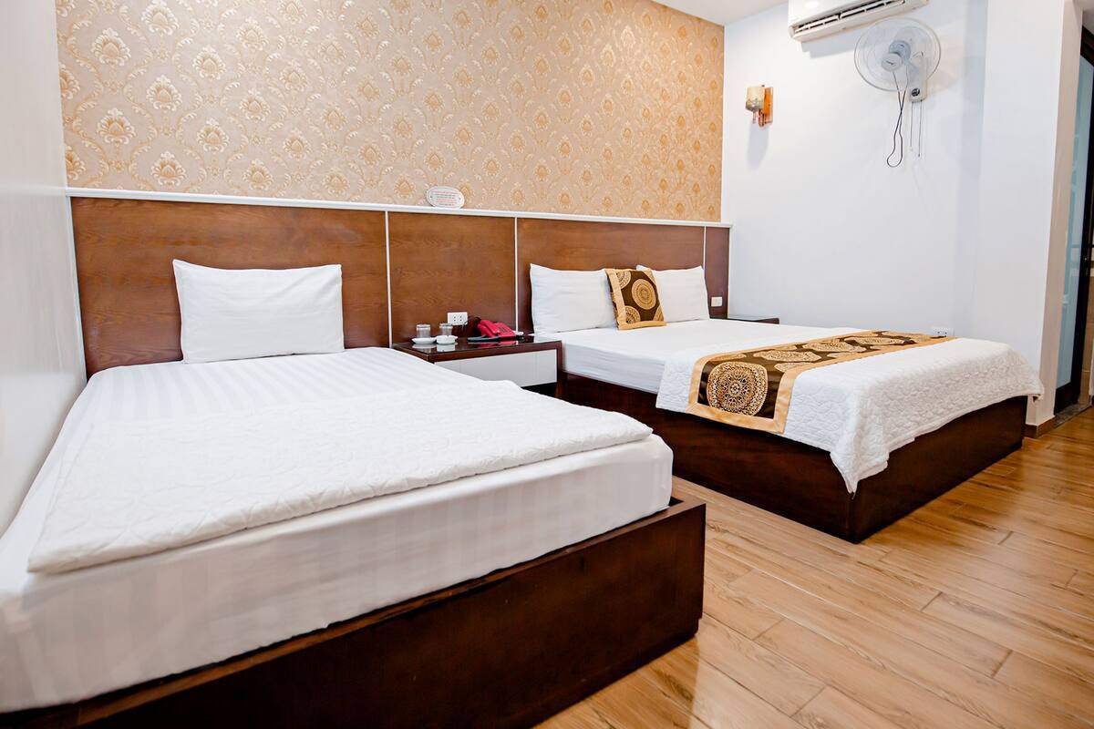 A hotel room with two beds, wooden headboards, a patterned wallpaper, an air conditioning unit, and a bedside table with a lamp.