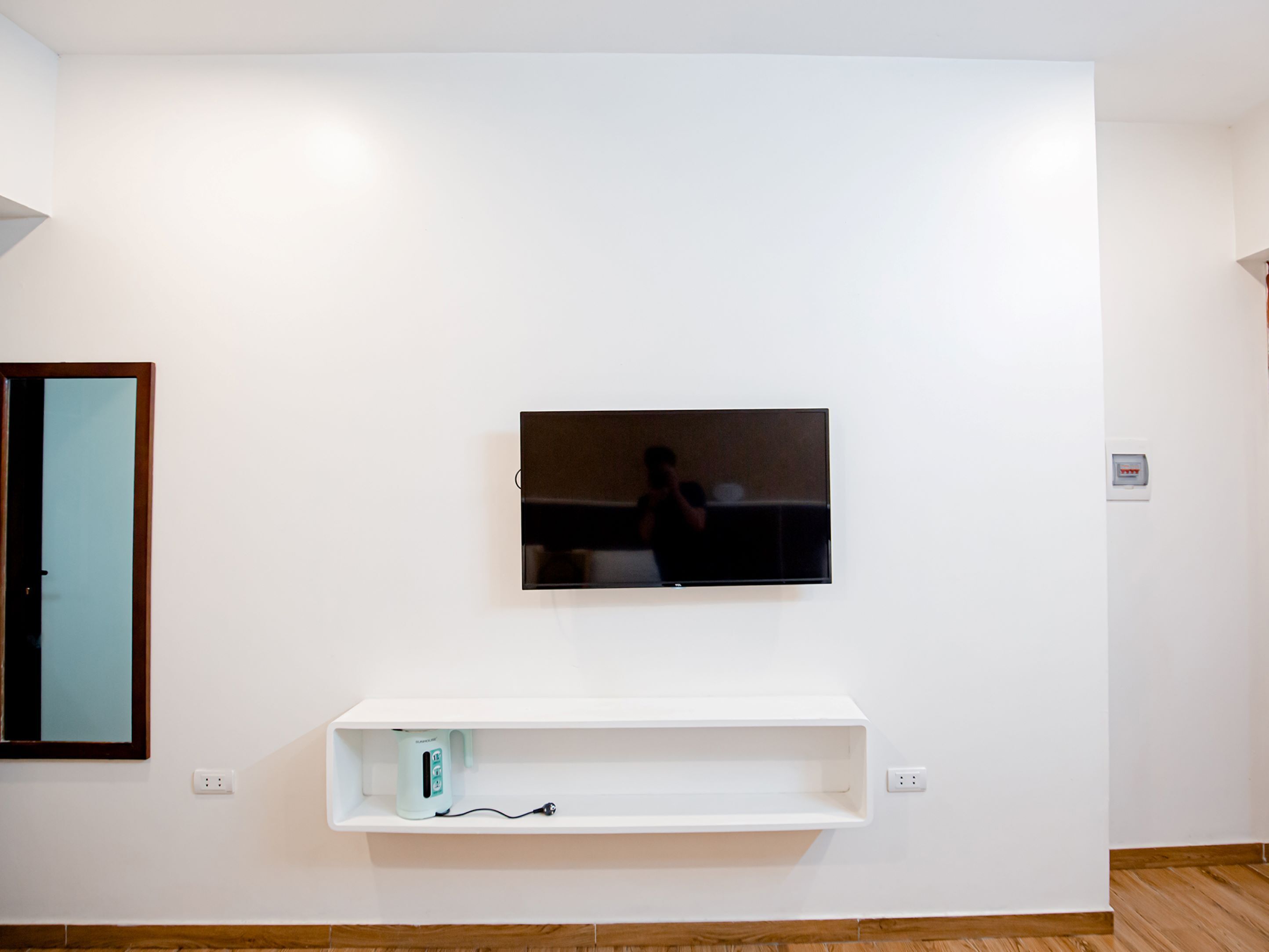 Une pièce équipée d’un téléviseur à écran plat fixé au mur, d’une étagère blanche située en dessous et d’une porte avec un miroir.