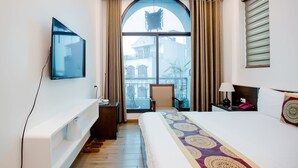 Habitación de hotel con una cama grande, televisor montado en la pared, escritorio con lámpara y vista a la ciudad a través de un ventanal amplio.