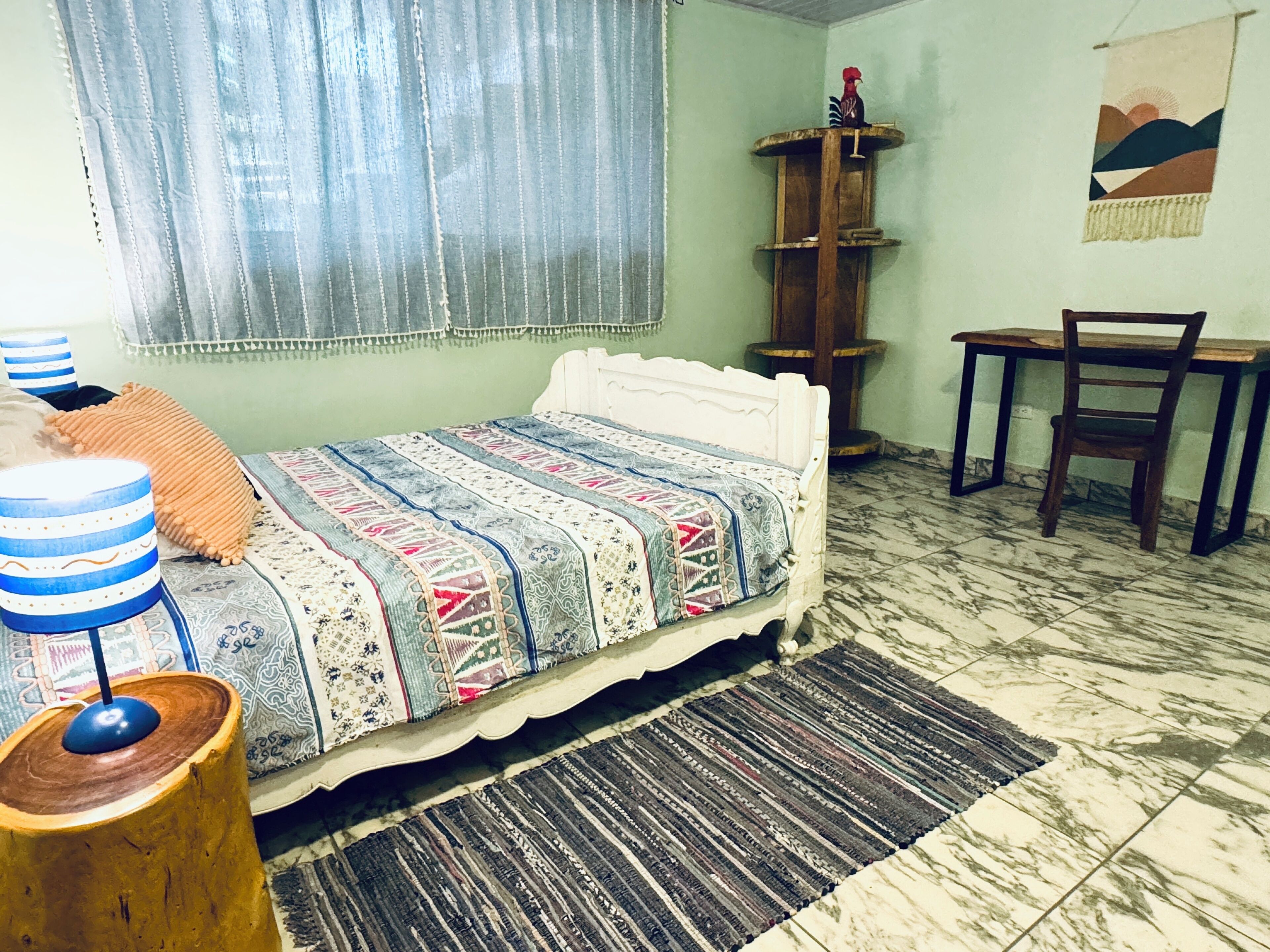 Ein Schlafzimmer mit einem Bett, einem Holzstuhl, einem kleinen Tisch, einem Holzschrank und einem Fenster mit Vorhängen.