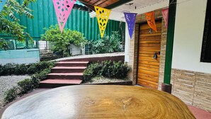 Ein Holztisch in einem Innenhof mit Stufen zu einer Tür, umgeben von Grünpflanzen und farbenfrohen dreieckigen Wimpeln.
