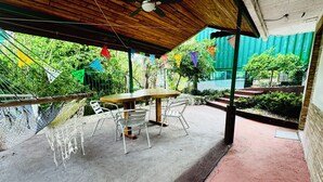 Eine Terrasse im Freien mit einem Holztisch und Metallstühlen, umgeben von einem Betonboden und einem grünen Metallzaun.