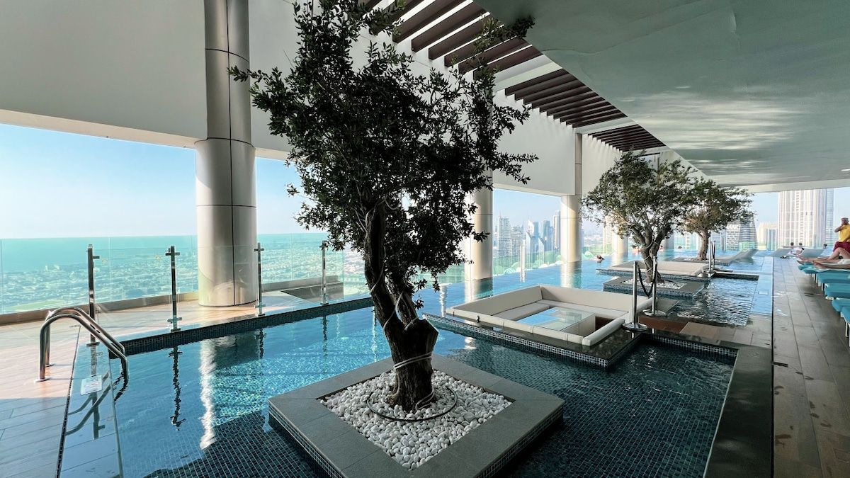 An indoor infinity pool with a tree centerpiece, lounge areas, and a view of the city skyline.