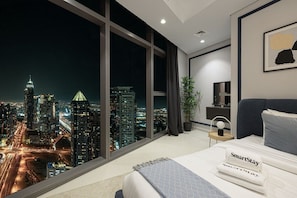 Ein modernes Hotelzimmer mit großem Fenster und Blick auf die Stadt, einem ordentlich bezogenen Bett und einem Nachttisch mit Lampe.