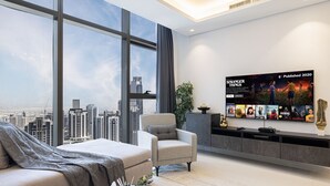 A modern living room with a large TV displaying 'Stranger Things' posters, a comfortable sofa, a grey armchair, and a city view through large windows. - 2BR |Sea & City Views |Infinity Pool |High Floor (Dubai)