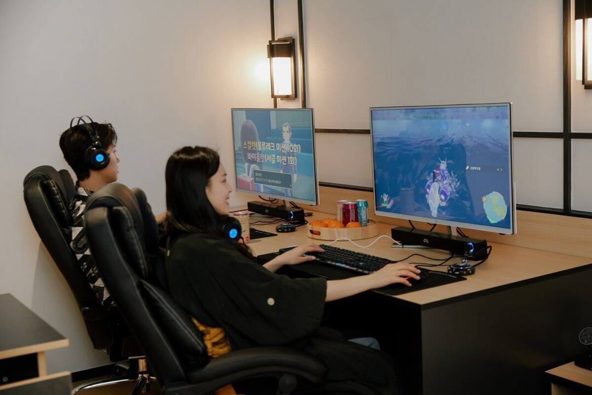Two individuals seated at a desk with a computer, headphones, and a monitor displaying a game.