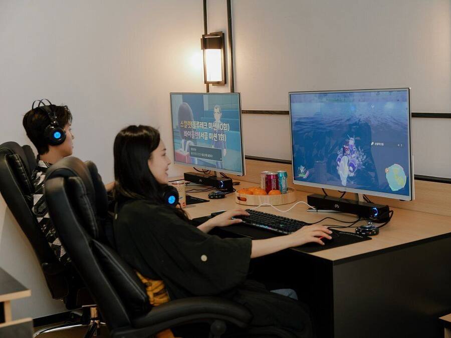Two individuals seated at a desk with a computer, headphones, and a monitor displaying a game.
