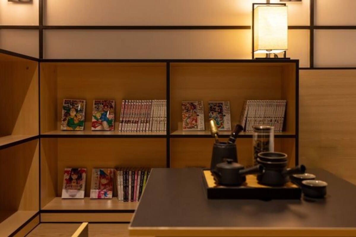 Una habitación con una estantería de madera que muestra varios libros y revistas, una mesa con una tetera y tazas, y una lámpara en la pared.