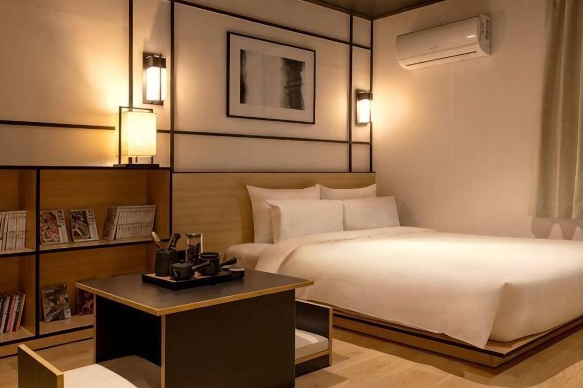 Quarto de hotel com cama, mesa de cabeceira, ar-condicionado fixado na parede e uma estante com livros.