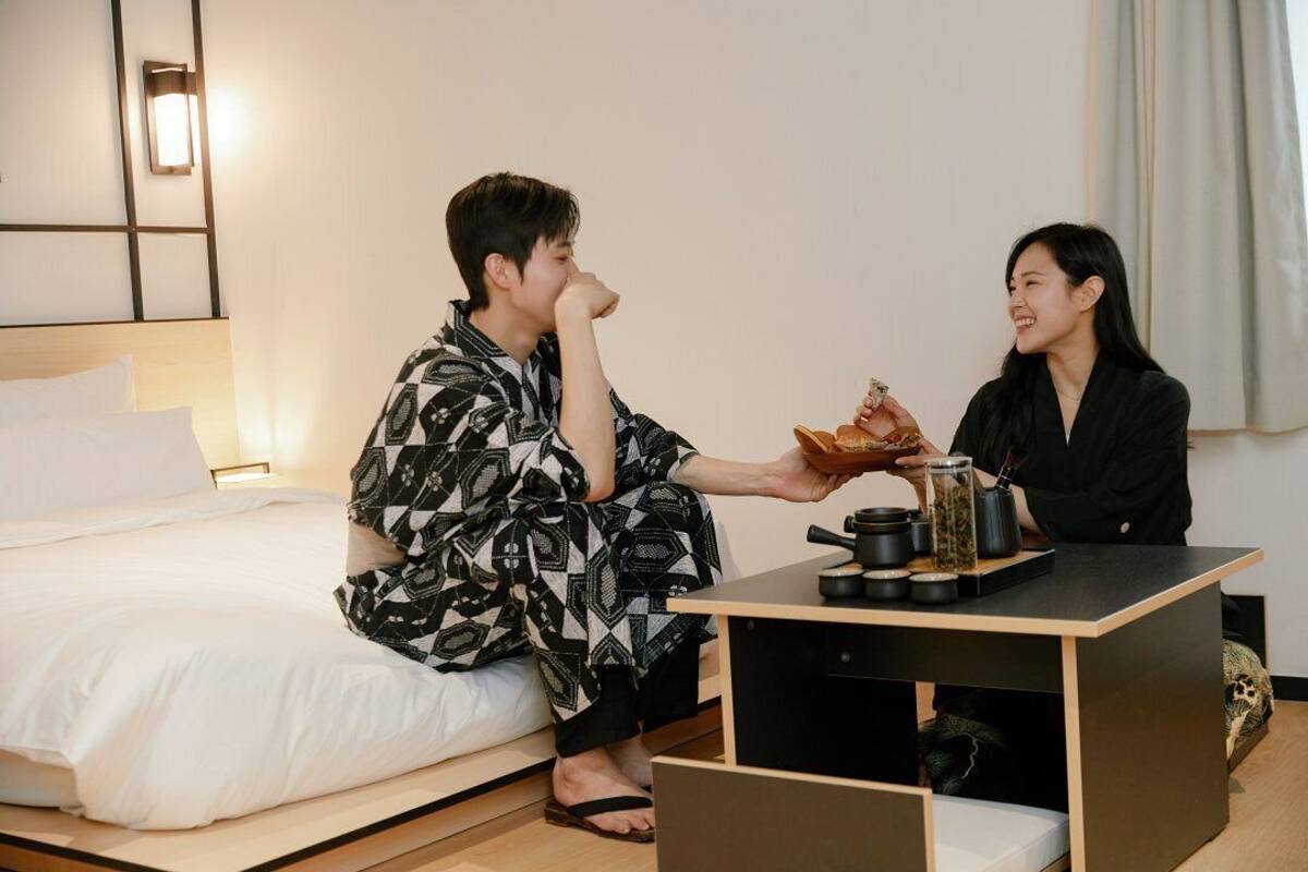 A room with a bed, a small table with jars and a cup, and a wall-mounted light.