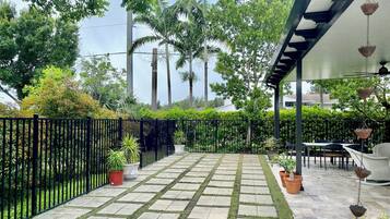 A paved patio with a black metal fence, potted plants, and outdoor furniture under a pergola.